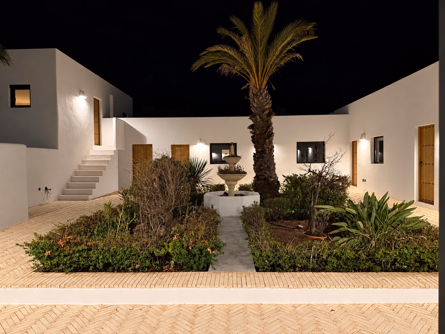 Patio la nuit : un palmier et une fontaine entre des murs blancs éclairés
