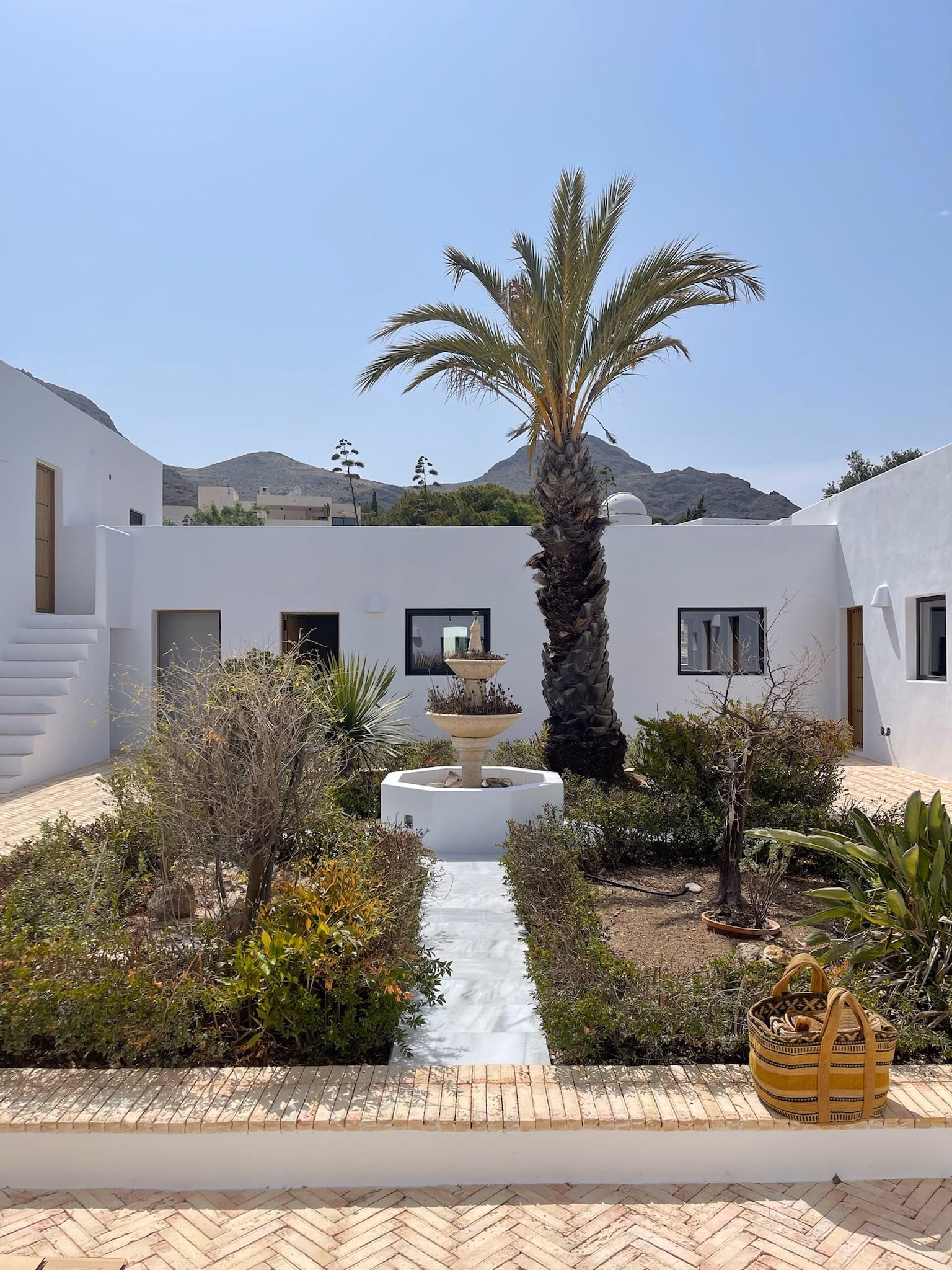 Patio blanc de jour, avec palmier, fontaine et massifs plantés, des collines au loin