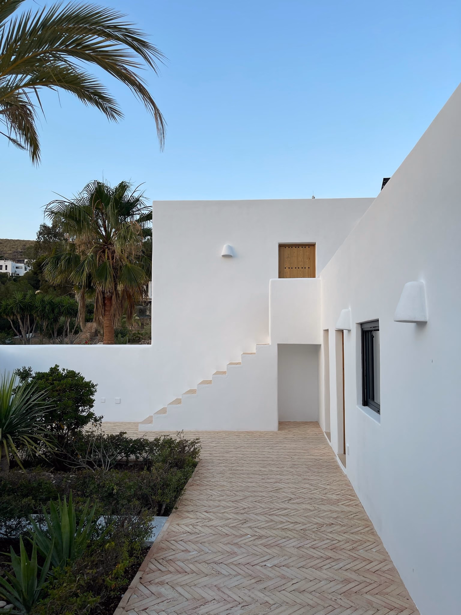Allée pavée entre des murs blancs, avec un escalier extérieur et des palmiers