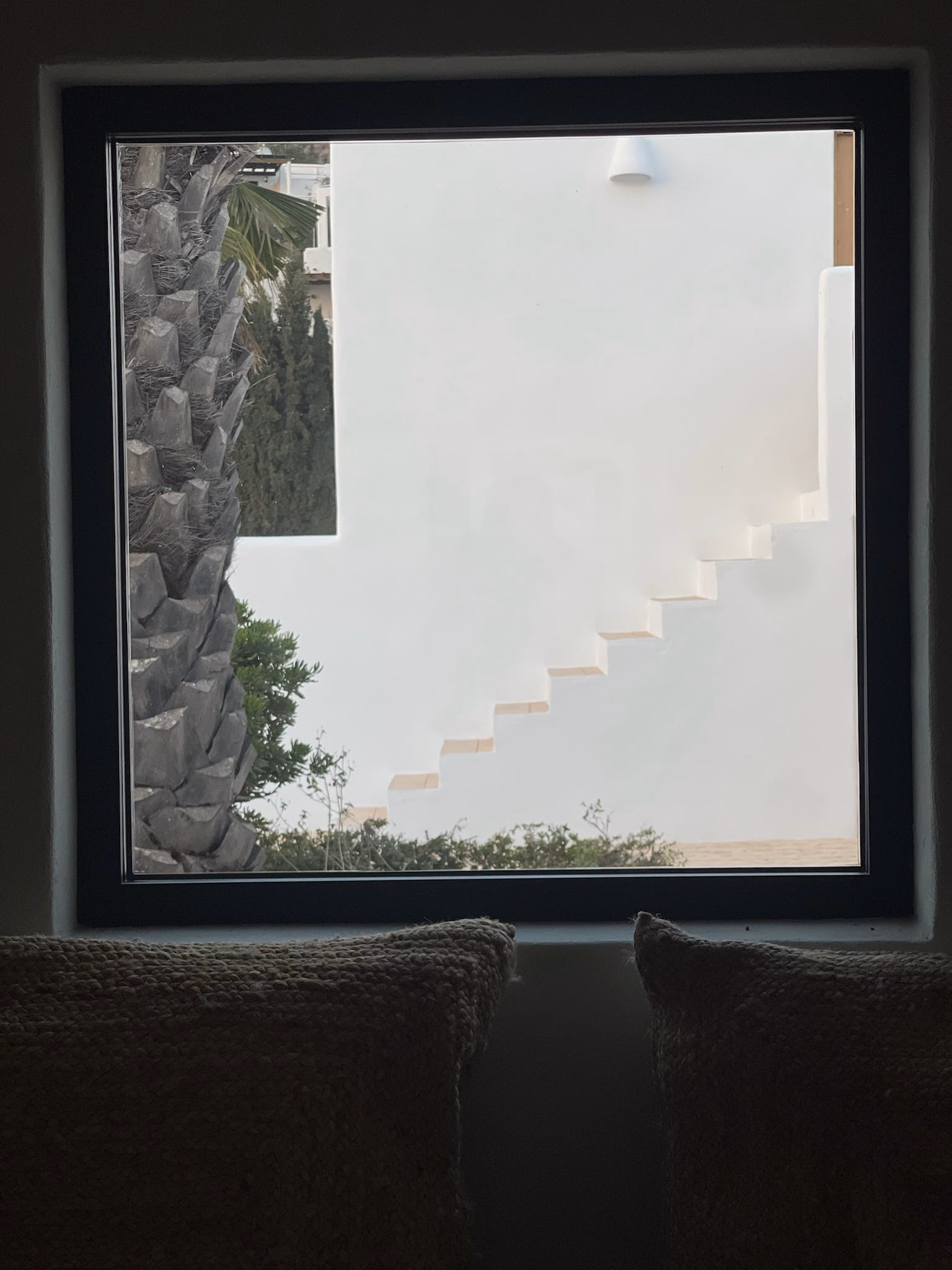 Fenêtre carrée qui cadre un mur blanc et son escalier extérieur