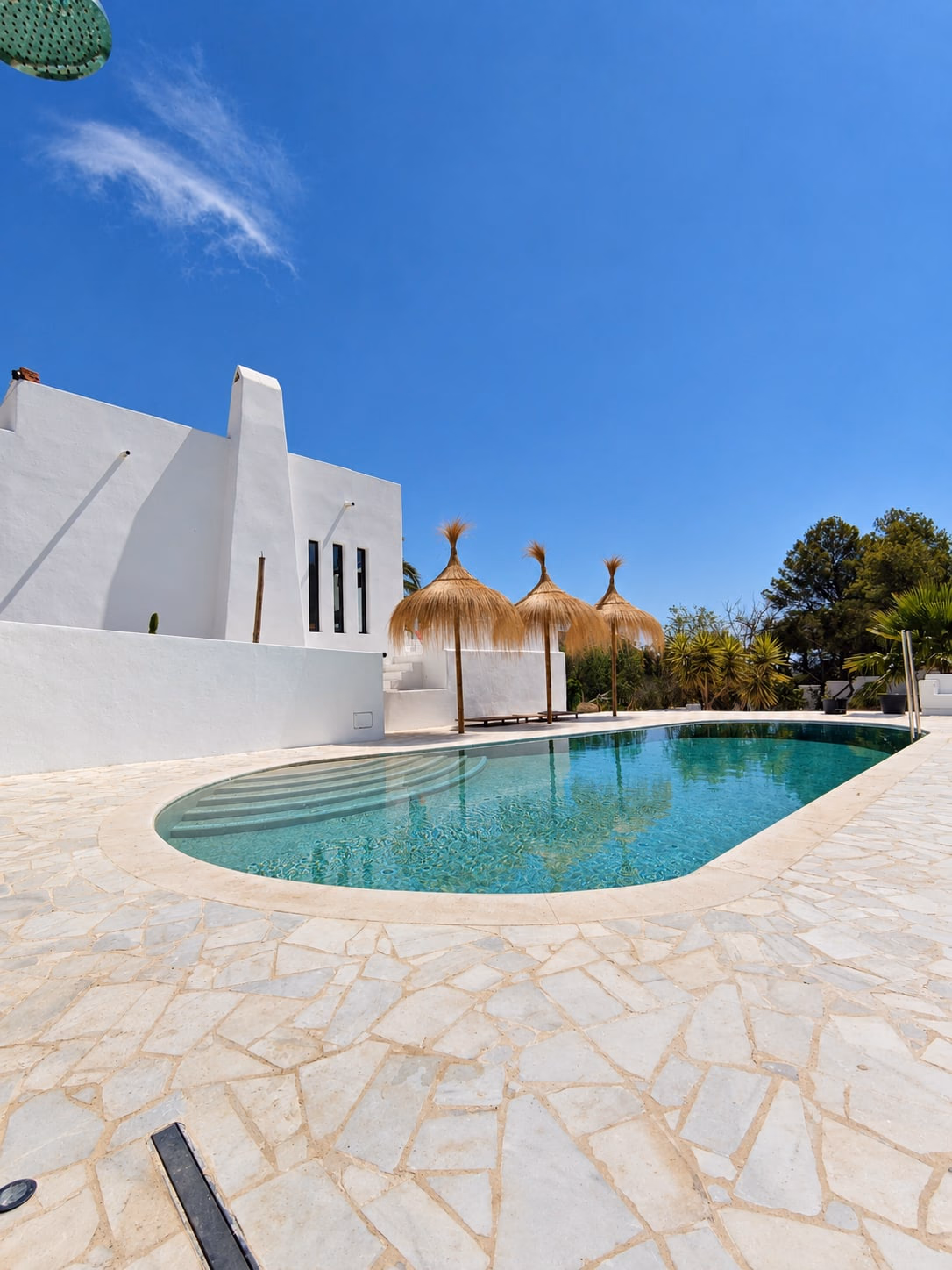 Maison blanche au bord d’une piscine ovale, parasols en paille et terrasse dallée