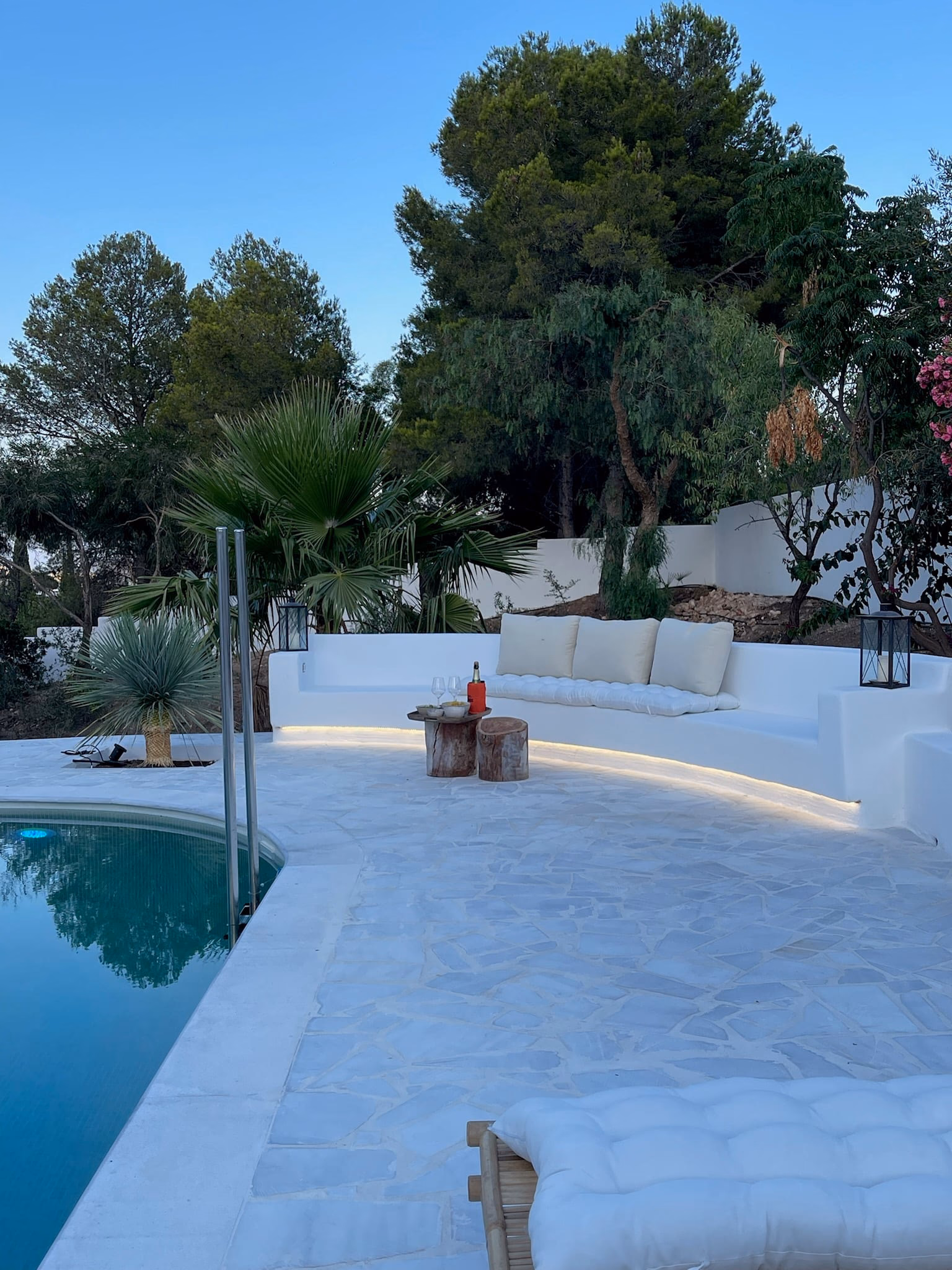 Piscine aux bords arrondis à côté d’une banquette blanche maçonnée avec coussins, des arbres derrière