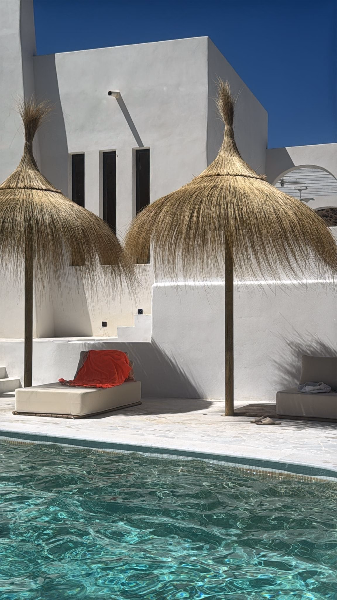 Parasols en paille sur une terrasse blanche au bord de la piscine, des murs blancs derrière