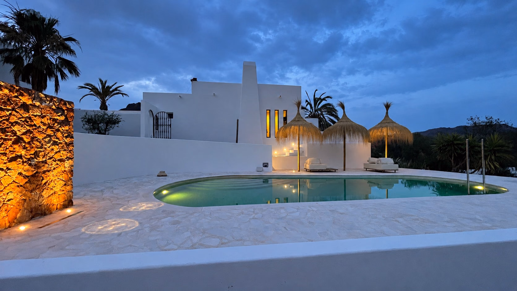 La piscine au crépuscule, parasols en paille et maison blanche au-delà, un mur de pierre éclairé
