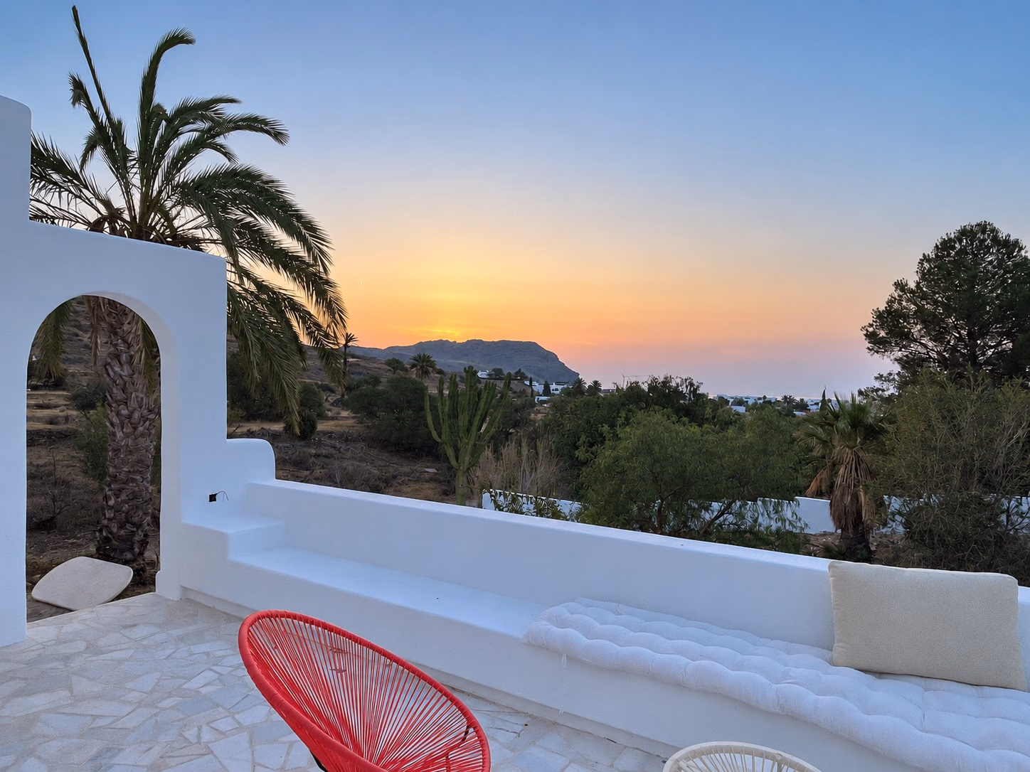 Terrasse au coucher du soleil avec banquette maçonnée et chaise rouge, une arche et des palmiers