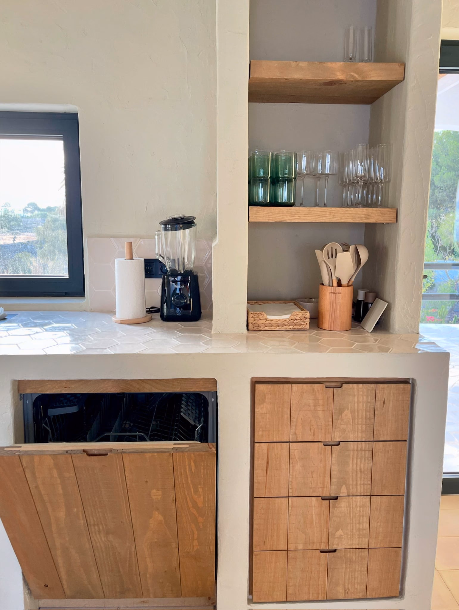 Détail de cuisine : tiroirs en bois sous un plan de travail blanc, verres sur des étagères ouvertes en bois